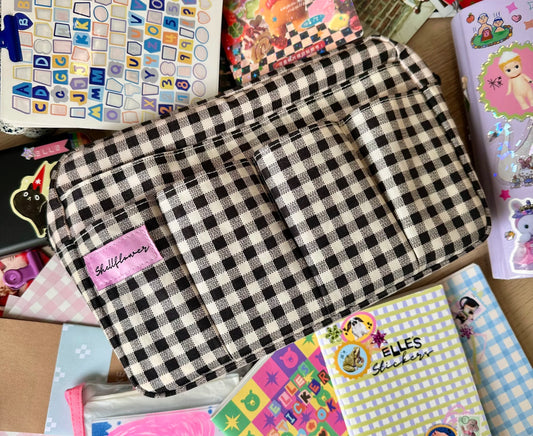 Black and white checkered pouch with 'Shelley's' label on a background of colorful stickers and paper.