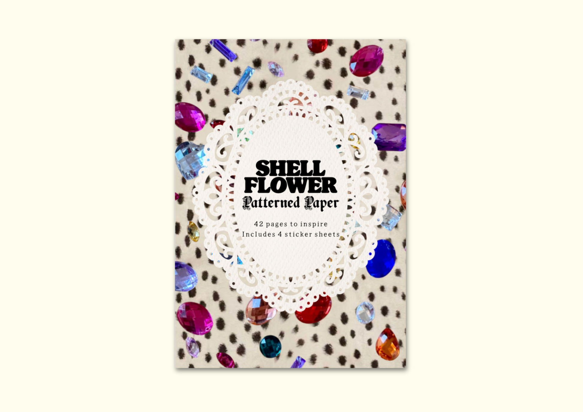 A book or booklet titled 'SHELL FLOWER Patterned Paper' shown upright on a light cream background. The cover features a white lace-like circular motif over a beige surface with scattered brown spots and colorful shell-like gem illustrations in red, blue, purple, and orange.