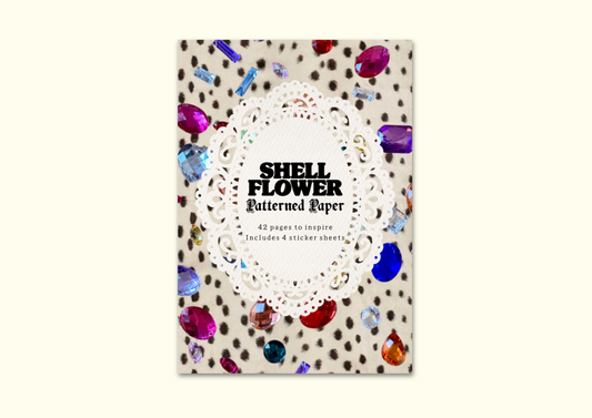 A book or booklet titled 'SHELL FLOWER Patterned Paper' shown upright on a light cream background. The cover features a white lace-like circular motif over a beige surface with scattered brown spots and colorful shell-like gem illustrations in red, blue, purple, and orange.
